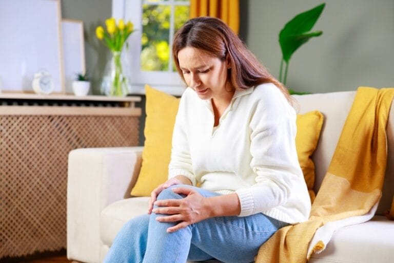 Mulher segurando o joelho sentada em um sofá branco com almofadas amarelas