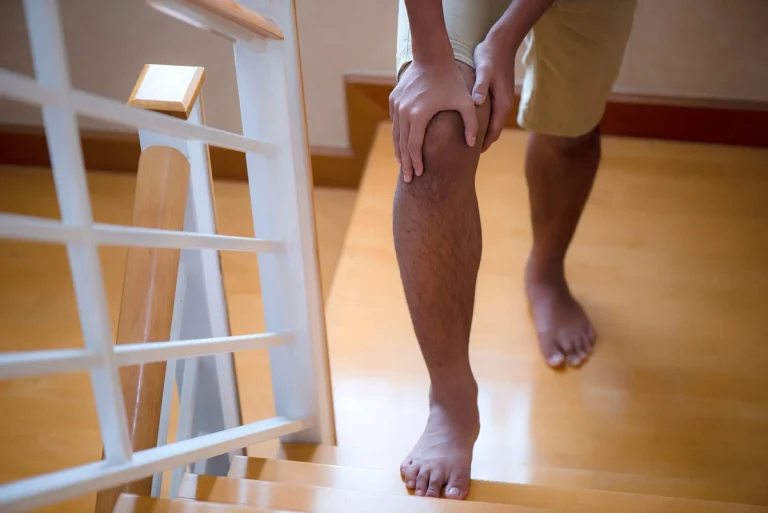 Homem subindo uma escada com a mão apoiada no joelho, demonstrando sentir dor e desconforto na articulação durante o esforço.